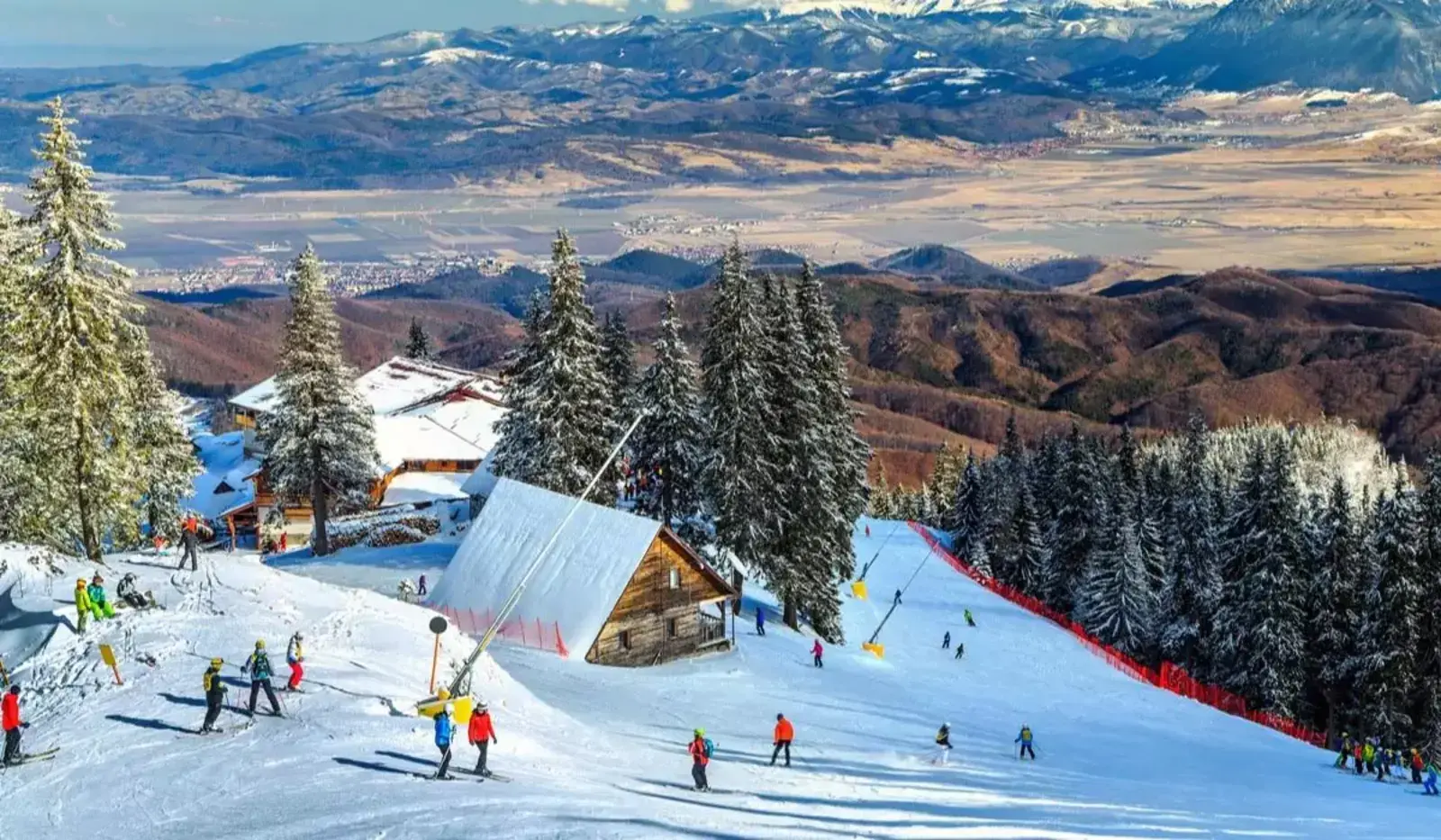 Schiatori pe p&acirc;rtie &icirc;n Poiana Brașov, munți și brazi &icirc;n fundal.